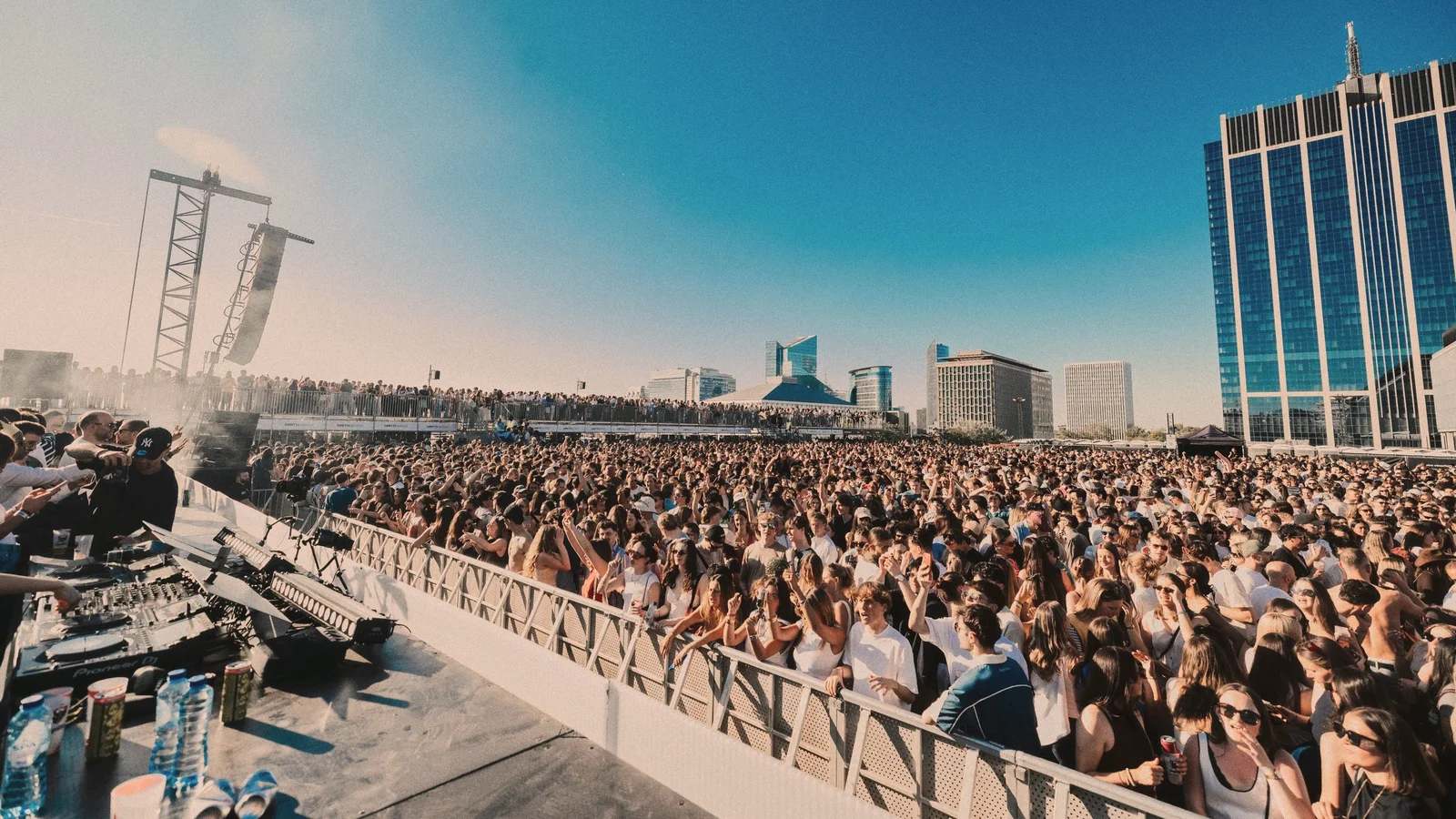 A sunlit open-air crowd in front of a stage, city towers behind